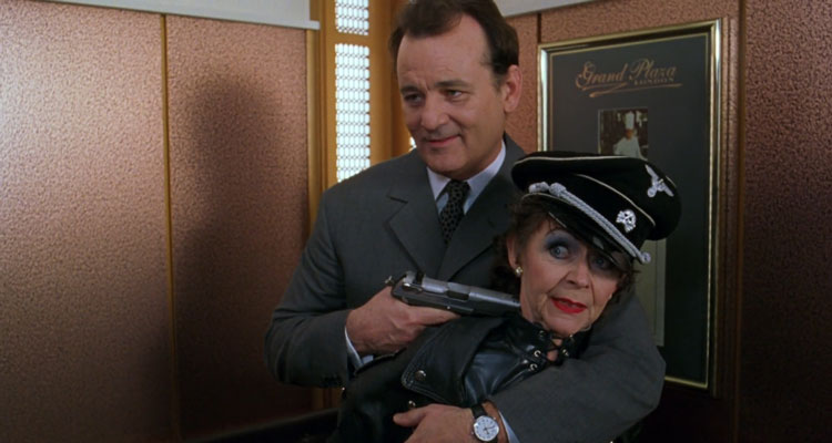 The Man Who Knew Too Little 1997 Movie Bill Murray as Wallace Ritchie holding a gun to an old lady dressed in leather with nazi cap