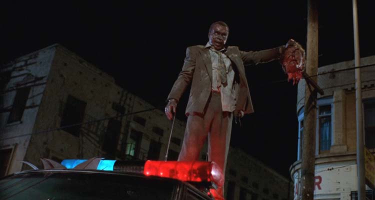 Tales From The Hood 1995 Movie Scene Tom Wright as Zombie Martin Moorehouse holding the severed head of Wings Hauser as Strom and standing on top of a police car
