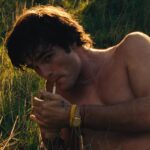 Saltburn 2023 Movie Scene Jacob Elordi as Felix Catton with his shirt off lighting a cigarette in a field