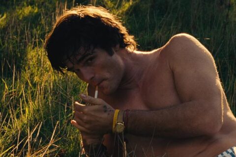 Saltburn 2023 Movie Scene Jacob Elordi as Felix Catton with his shirt off lighting a cigarette in a field