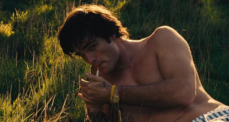 Saltburn 2023 Movie Scene Jacob Elordi as Felix Catton with his shirt off lighting a cigarette in a field