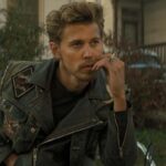 The Bikeriders 2023 Movie Austin Butler as Benny smoking a cigarette and wearing a leather jacket while sitting on his motorcycle