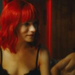 Strange Darling 2023 Movie Scene Willa Fitzgerald as The Lady in a bra and red wig sitting on a bed of a motel room
