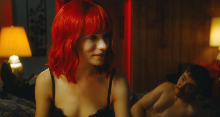 Strange Darling 2023 Movie Scene Willa Fitzgerald as The Lady in a bra and red wig sitting on a bed of a motel room