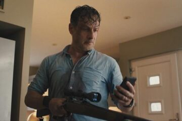 Coldwater 2025 Series Scene Andrew Lincoln as John looking at the phone while holding a rifle